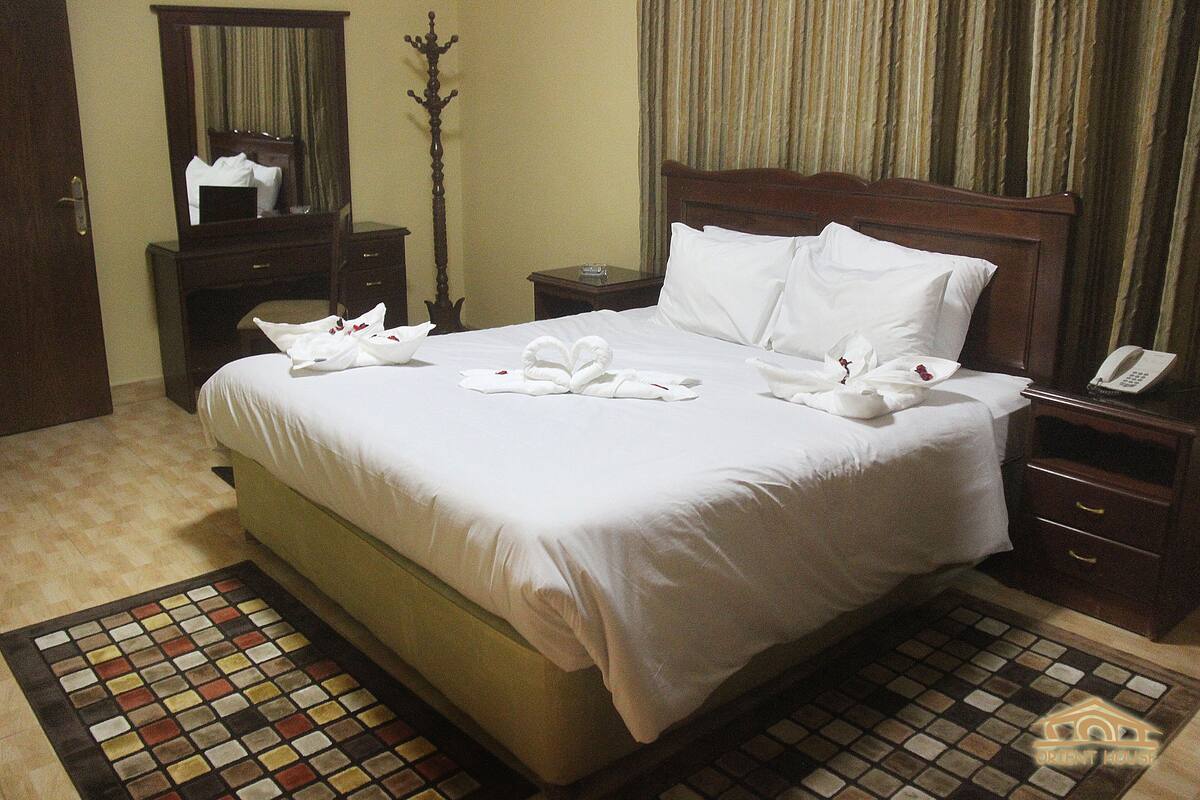 A neatly made bed with white linens and towels arranged in a heart shape, a wooden nightstand with a phone, and a mirror above the dresser.