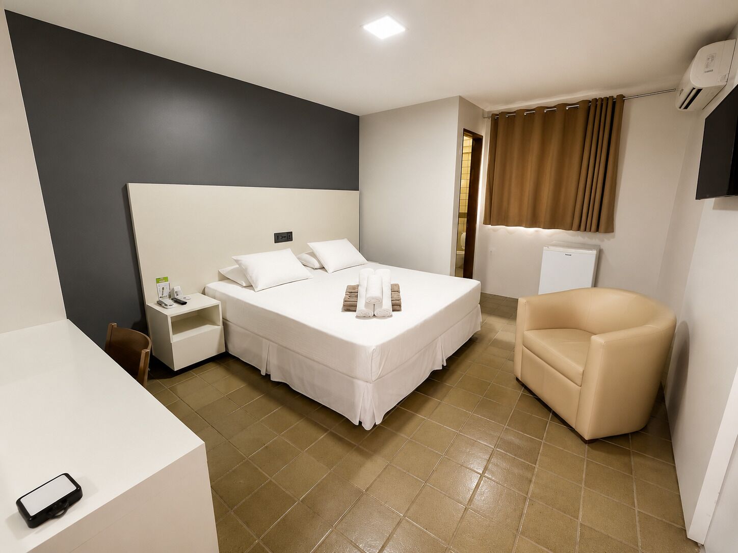 Quarto de hotel com cama, mesa de cabeceira, cadeira e uma pequena escrivaninha com telefone.