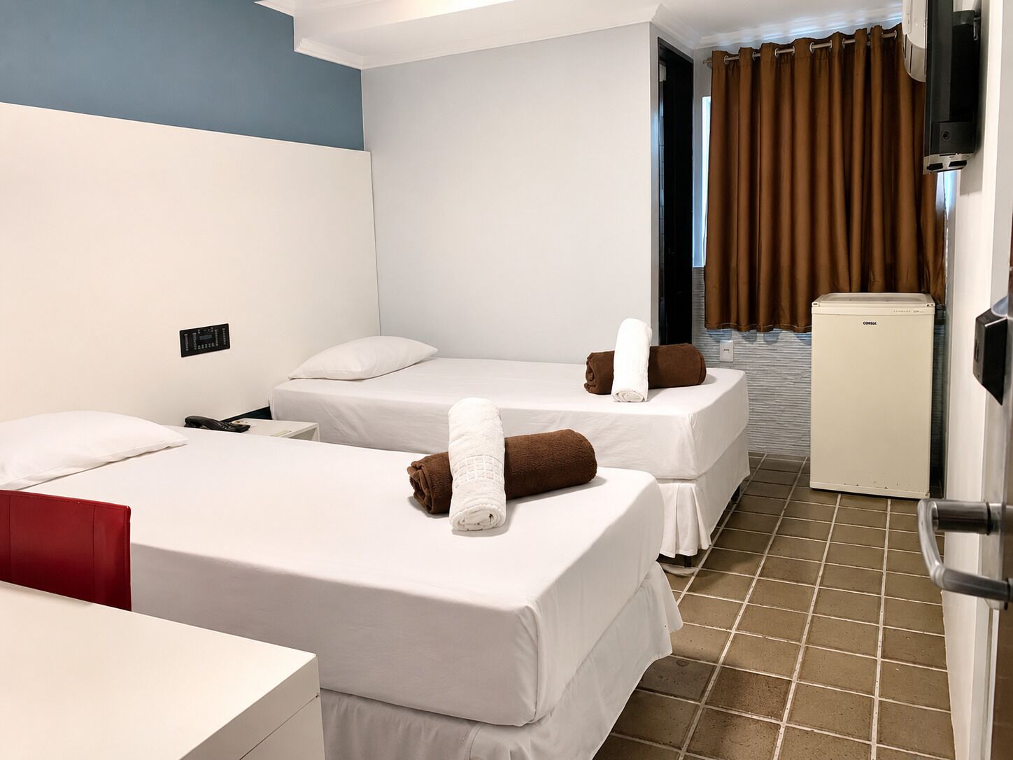 A hotel room with two single beds, a small refrigerator, a desk with a chair, and a bathroom visible through an open door.