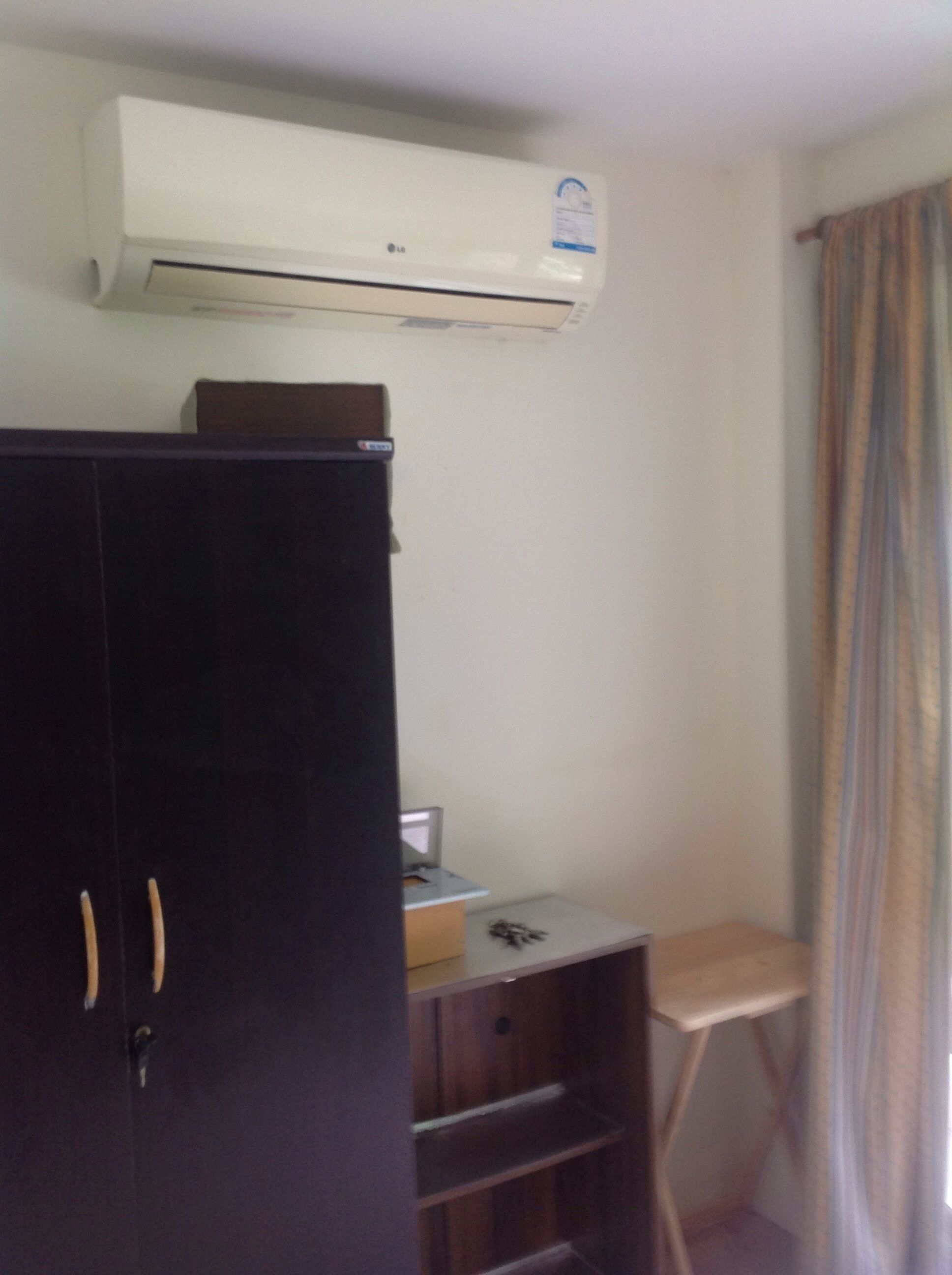 A room with a dark wooden wardrobe, a small desk with a chair, and an air conditioning unit mounted on the wall.