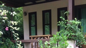A covered porch with a wooden bench, surrounded by lush greenery and flowering plants.