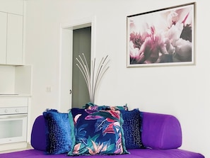 A modern living room with a purple sofa, a floral throw pillow, and a framed abstract painting.