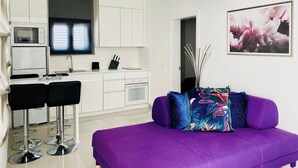 Un salon moderne avec un canapé violet, un téléviseur et un coin cuisine doté de meubles blancs.