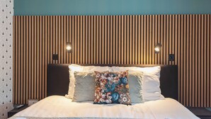 Un lit bien fait, agrémenté d’un coussin à motifs floraux, une tête de lit avec des lattes de bois verticales et deux lampes fixées au mur.