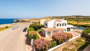 Una propiedad costera con una casa grande, piscina y una carretera con coches aparcados.