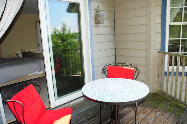 A small balcony with a round table and two red chairs, a sliding glass door, and a small bed.