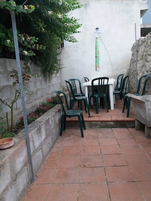 A patio area with a white table and green plastic chairs, surrounded by a low brick wall and a concrete planter with red flowers.