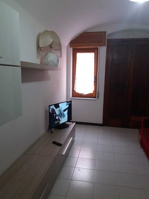A room with a television on a stand, a window with a curtain, a wooden door, and a shelf with a decorative item.