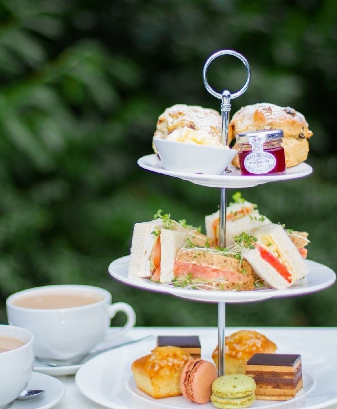 Etagebordsdisplay med sandwiches, scones og kager, ledsaget af kopper te og kaffe.
