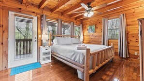 A wooden cabin bedroom with a bed, a nightstand, a ceiling fan, and a window with curtains. - Time to Get Cozy! King Bed I Hot Tub I Foosball Table I Cabin for Couples (Sevierville)