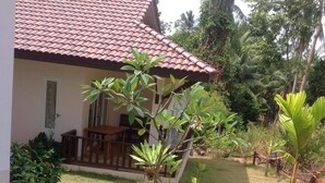 Ein Haus mit einem Ziegeldach, einer Veranda mit Tisch und Stühlen sowie ein kleiner Garten mit verschiedenen Pflanzen und einer Palme.