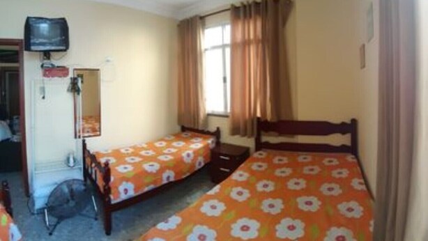 A room with two single beds, a ceiling fan, a television, and a window with curtains. - Apartment 170m from Praia do Forte (Centro)
