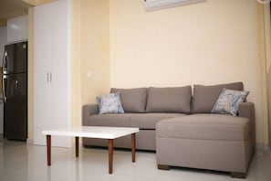 A living room with a sofa, two patterned pillows, a white coffee table, and a kitchen area with a refrigerator.