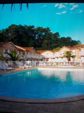 A swimming pool surrounded by white buildings with balconies, palm trees, and a clear blue sky.