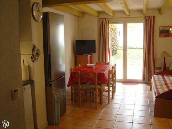 A compact kitchen with a refrigerator, a dining table with chairs, a television, and a bed with a checkered bedspread.