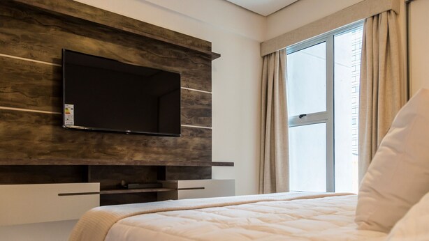 Ett modernt hotellrum med en stor säng, en platt-TV som är monterad på en väggpanel i trä och ett fönster med gardiner.