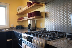 A kitchen with a gas stove, wooden shelves with dishes, a coffee maker, and a tiled backsplash. - Lake Michigan Waterview Family Retreat - Steps to Beach & Downtown Charlevoix (Charlevoix)