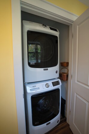A laundry room with a stacked washer and dryer, a basket, and a door. - Lake Michigan Waterview Family Retreat - Steps to Beach & Downtown Charlevoix (Charlevoix)