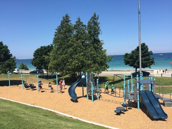 A playground with slides and swings, located near a beach with trees and a clear sky.
