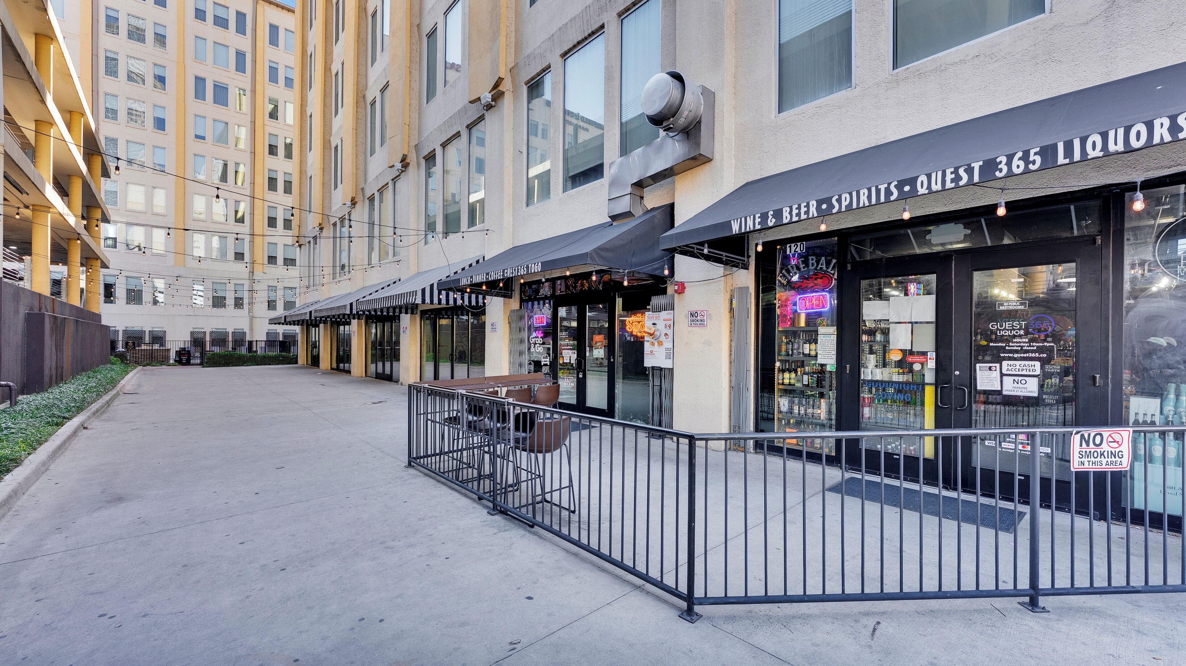 Une rangée de sièges extérieurs avec une rambarde en métal, une devanture de magasin avec une enseigne indiquant « Guest Liquor » et « Wine & Beer - Spirits - Quest 365 Liquors », et un bâtiment avec de nombreuses fenêtres en arrière-plan.