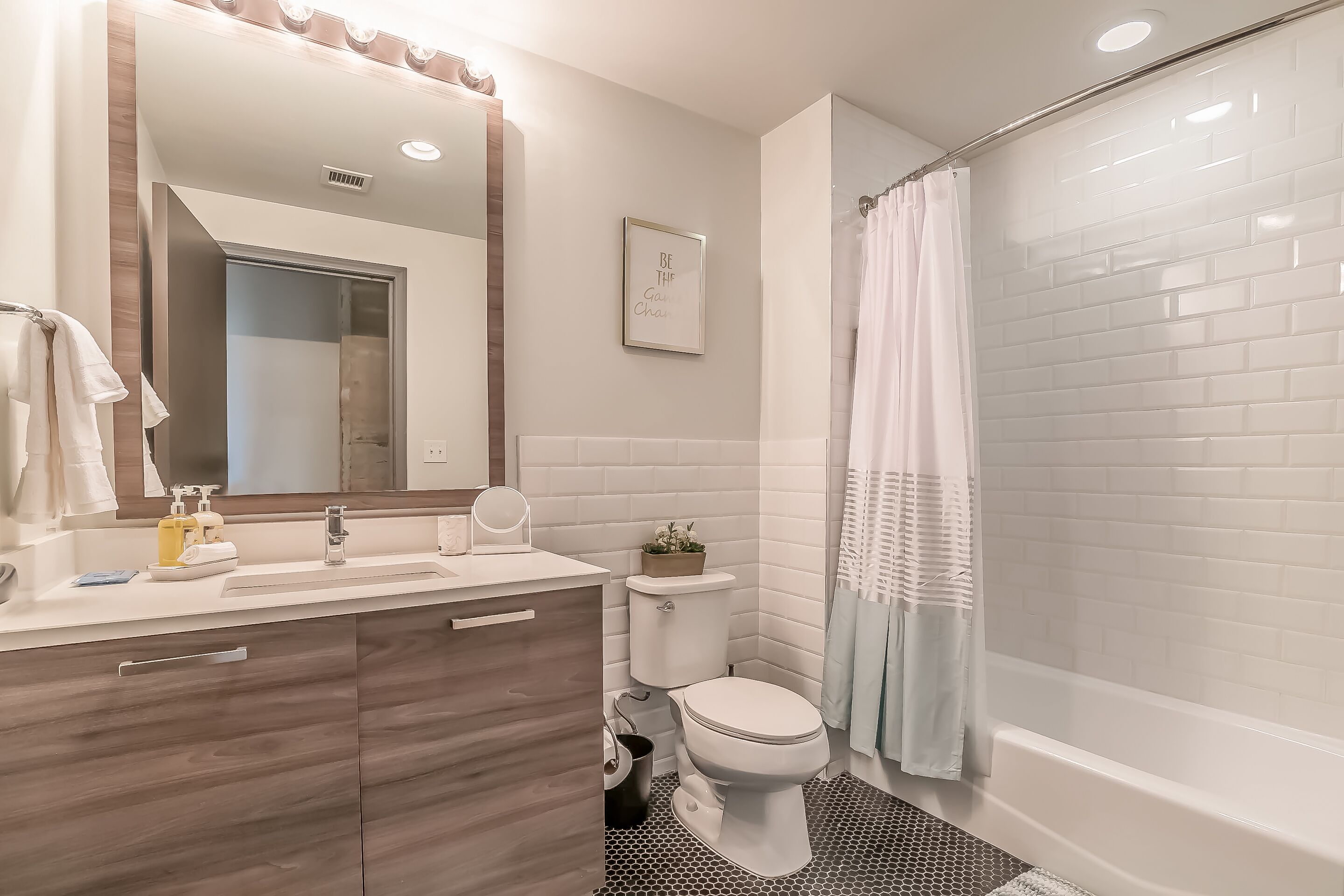Un baño moderno con un lavamanos, un espejo y un inodoro blancos, una ducha con cortina y una pequeña planta en maceta sobre el inodoro.