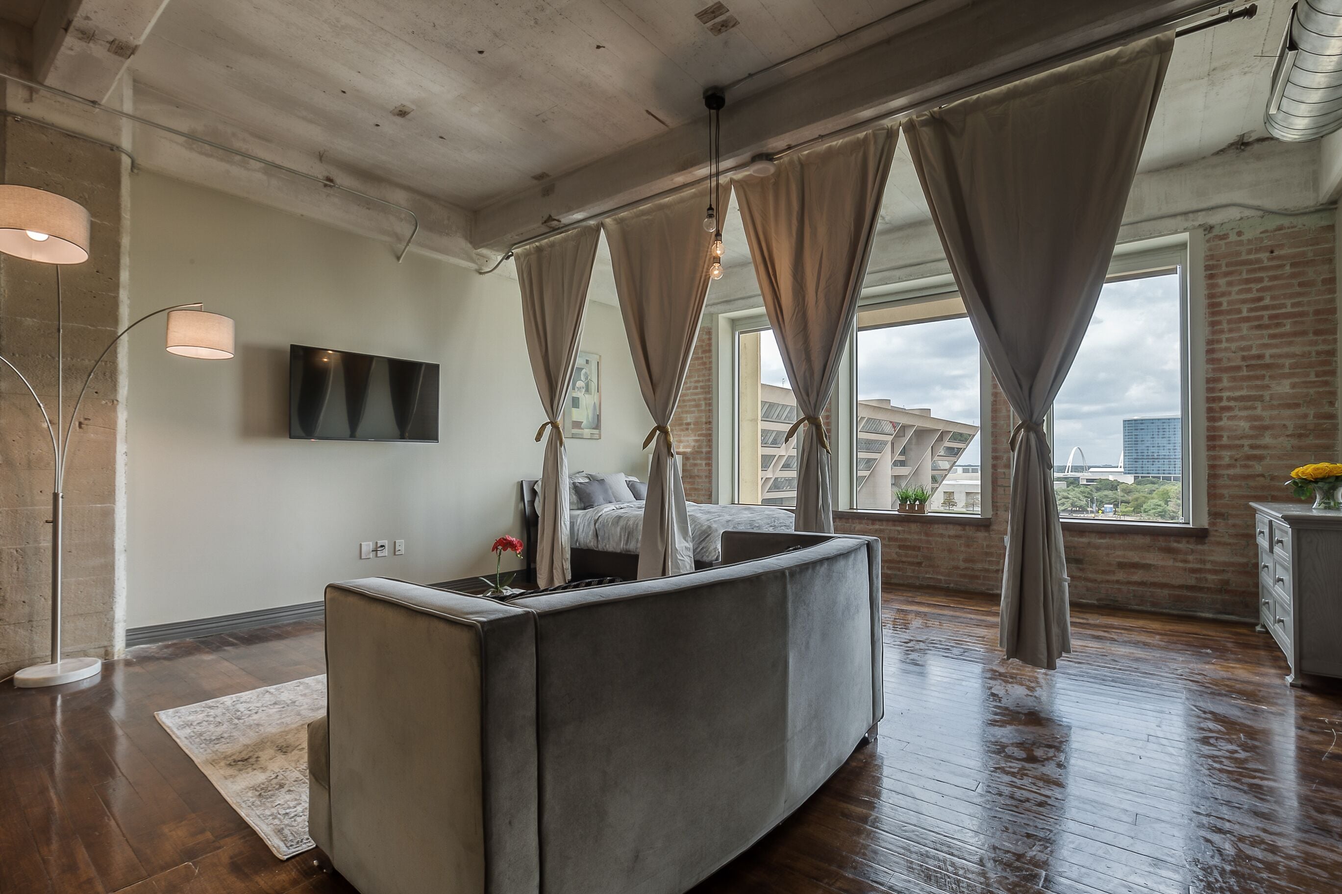 A modern bedroom with a large bed, a bedside table, a TV, and a view of the city through large windows.