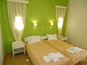 A hotel room with two beds, white curtains, bedside lamps, and a small table with a vase of flowers.