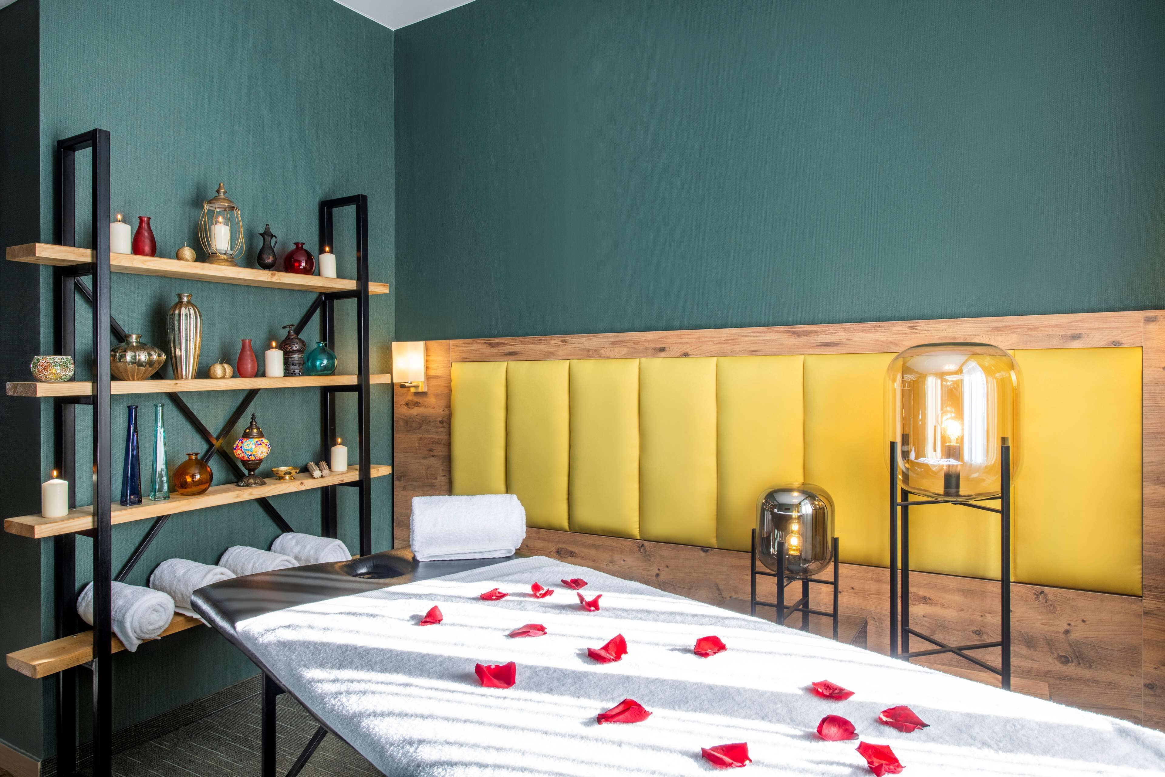 A spa room with a massage table, rose petals, shelves with decorative items, and a yellow padded headboard.