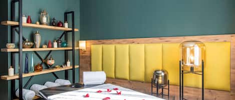 A spa room with a massage table, rose petals, shelves with decorative items, and a yellow padded headboard.