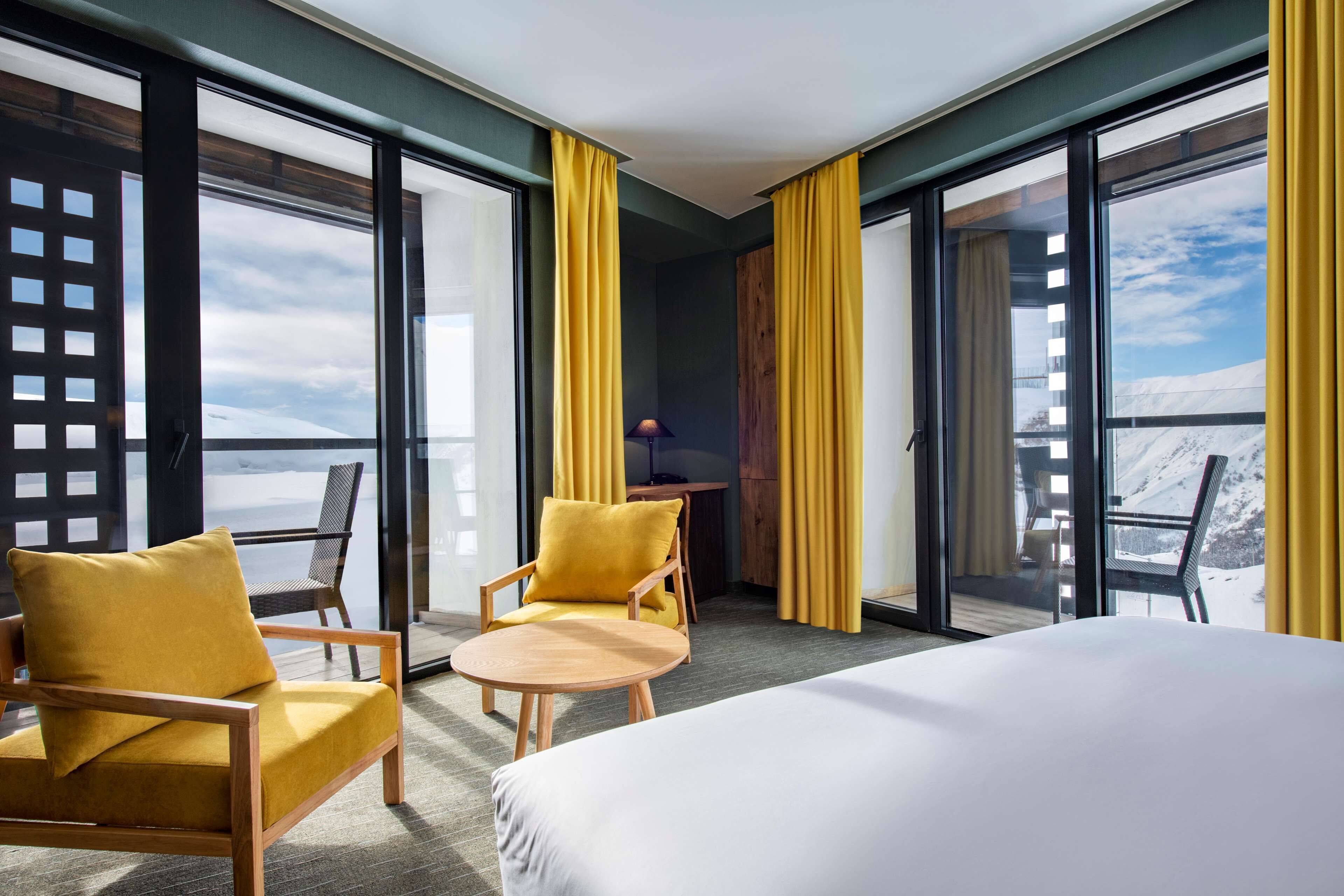Une chambre d’hôtel avec un lit, deux fauteuils jaunes, une petite table ronde et de grandes fenêtres donnant sur une vue sur les montagnes.