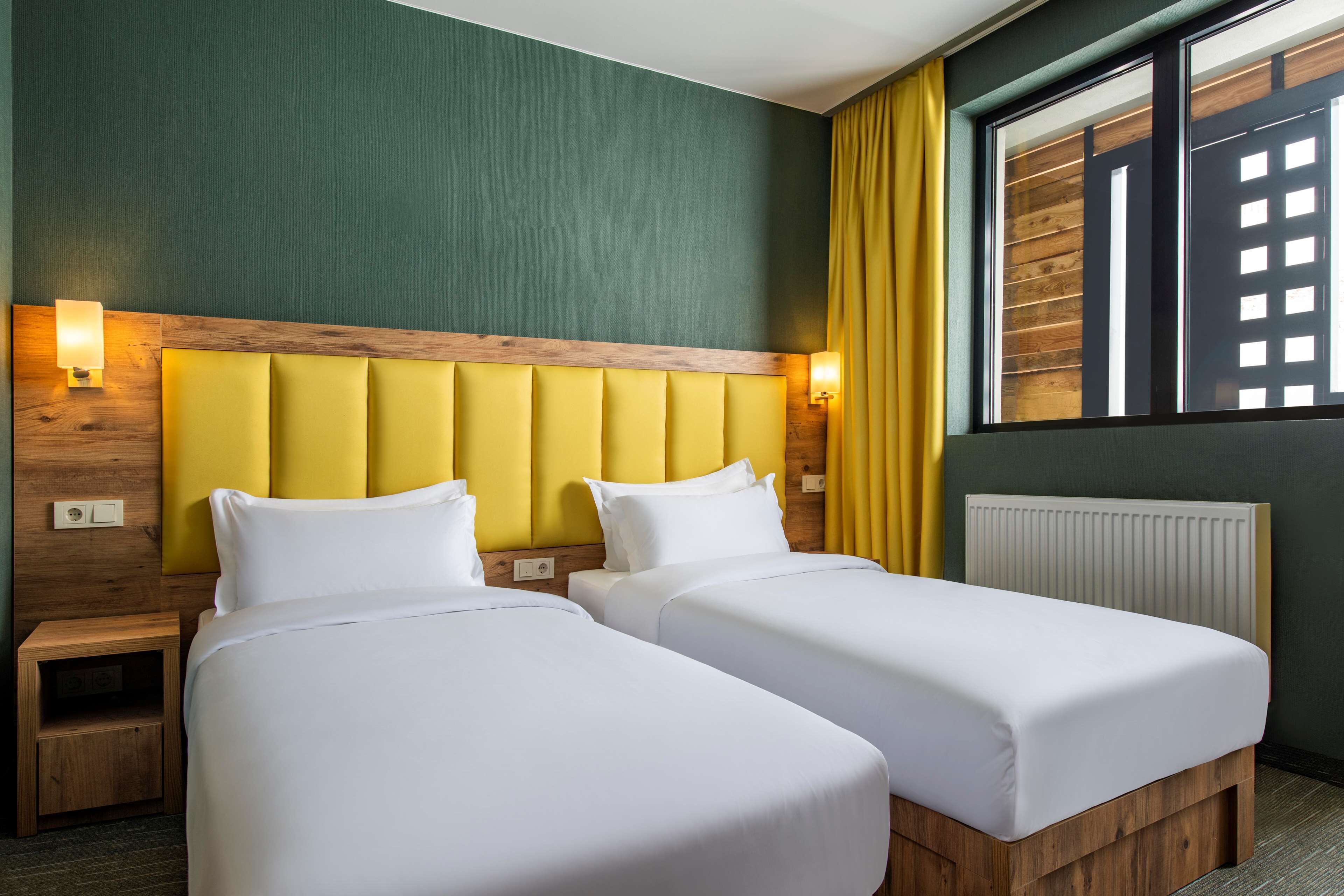 Une chambre d’hôtel avec deux lits, une tête de lit en bois, une table de chevet et une fenêtre avec des rideaux jaunes.