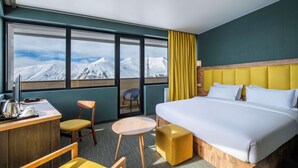 Habitación de hotel con una cama grande, un escritorio con silla, una mesita redonda y vistas a montañas nevadas a través de amplios ventanales.