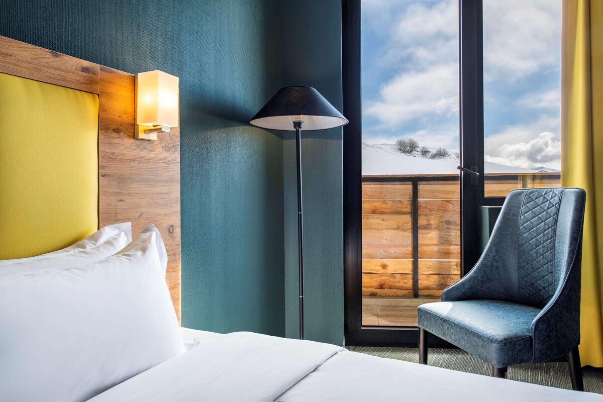 Una habitación de hotel con cama, una silla, una lámpara y vistas a un paisaje nevado a través de un ventanal grande.
