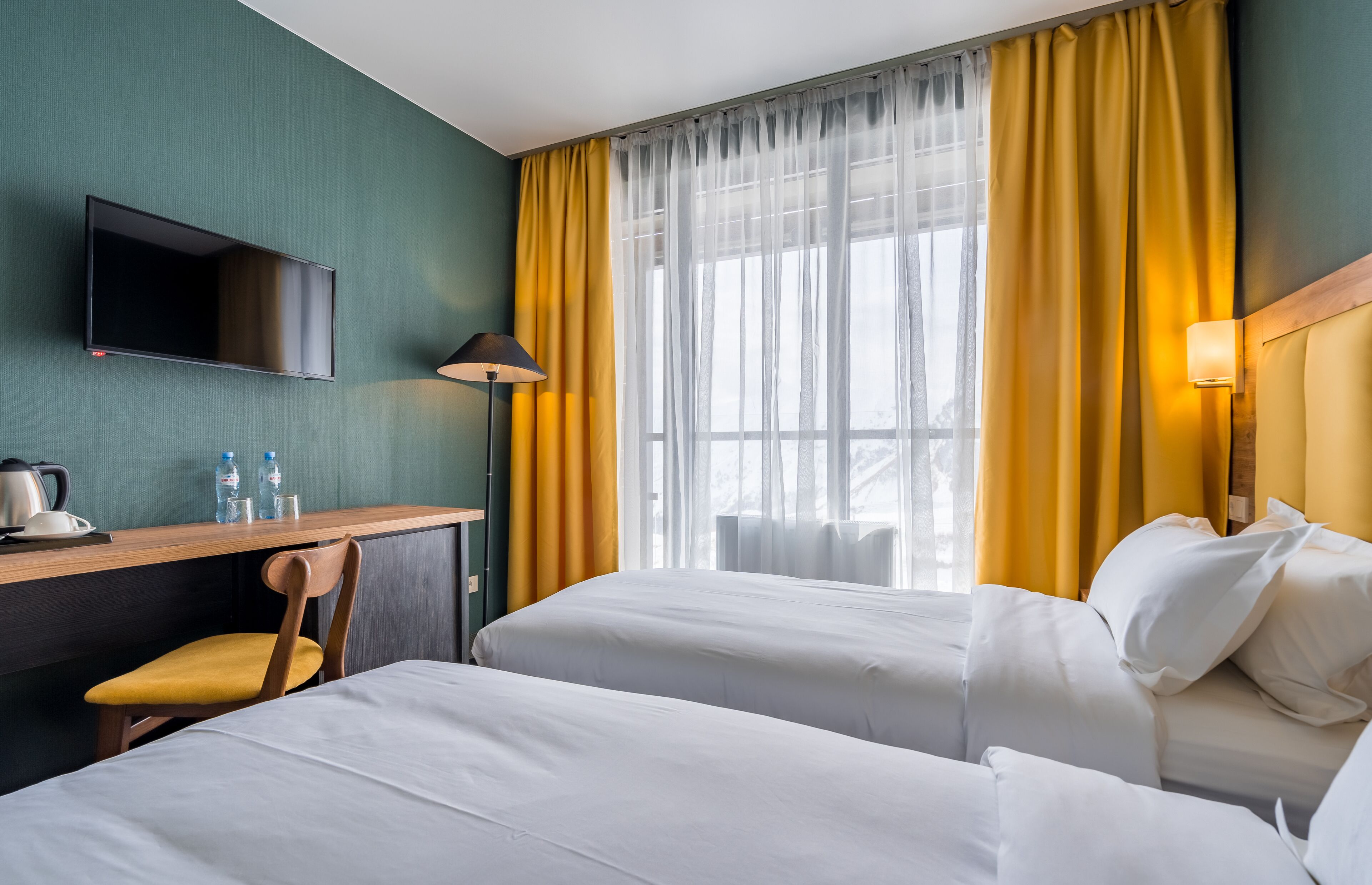Habitación de hotel con dos camas, un escritorio con silla, un televisor de pantalla plana, una ventana con cortinas amarillas y una lámpara.