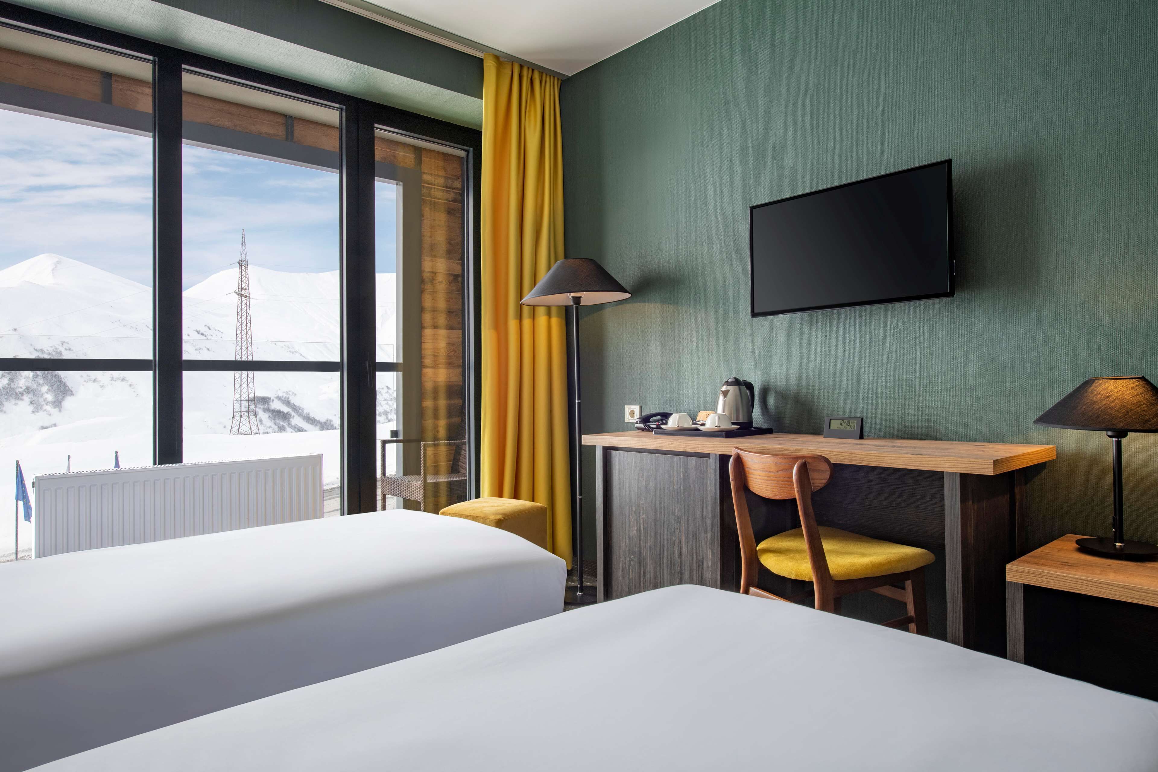A hotel room with two beds, a desk, a chair, a TV, and a view of snow-capped mountains.