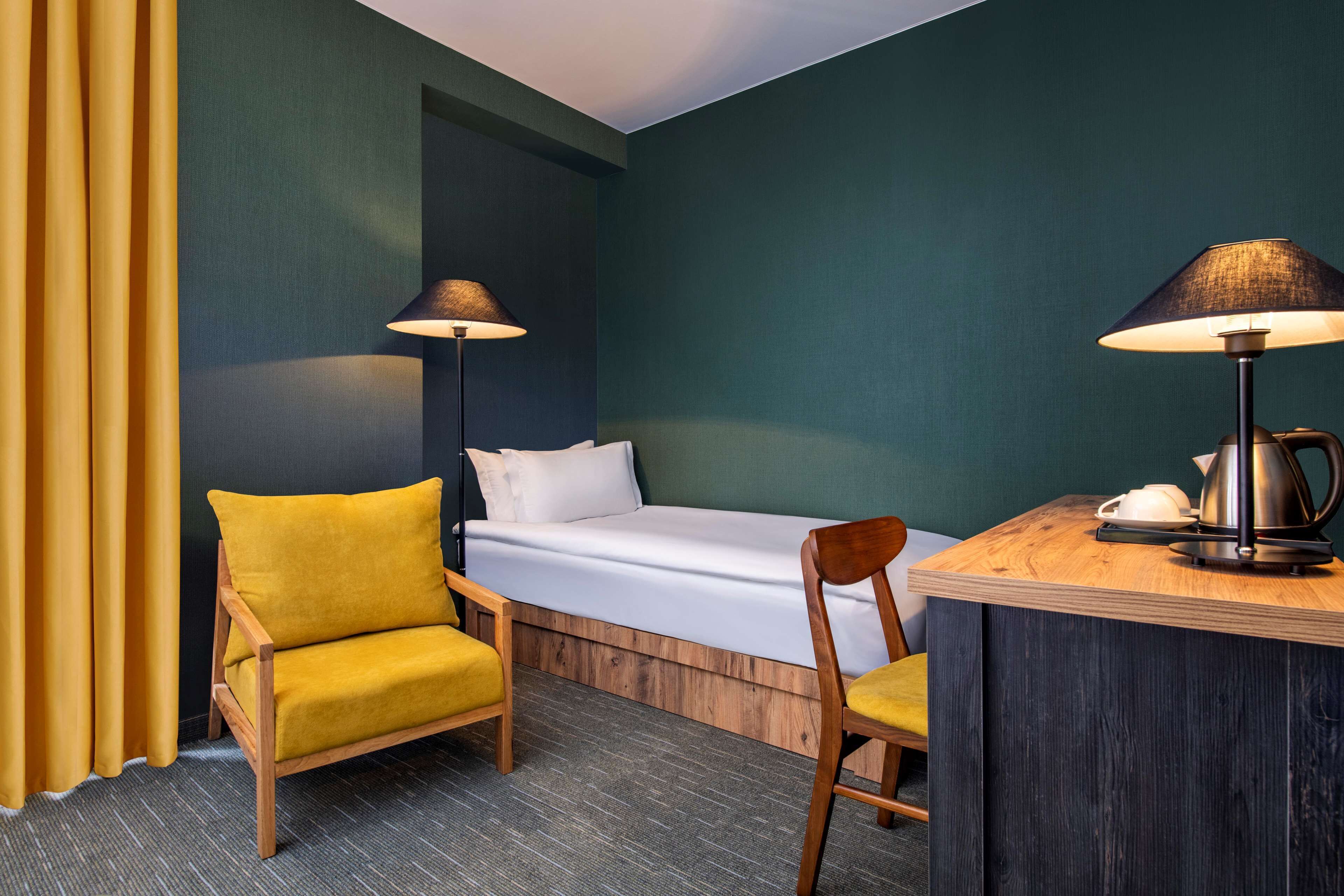 Une chambre d’hôtel de taille réduite, comprenant un lit simple, un bureau en bois, deux chaises et deux lampes.