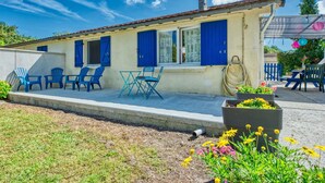 Ein kleines, eingeschossiges Gebäude mit blauen Fensterläden, einer Terrasse mit Sitzgelegenheiten und einem Gartenbereich mit Blumen.