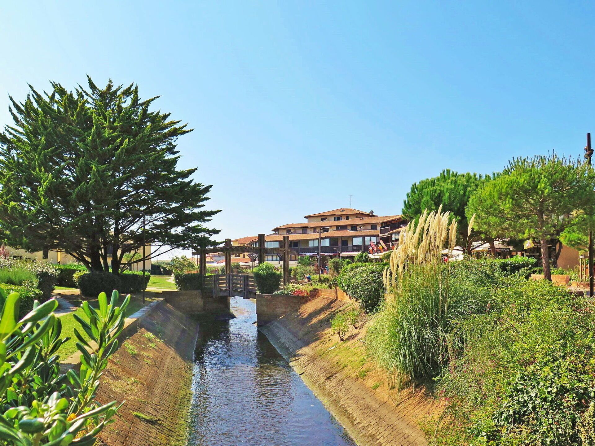 En kanal med en træbro, omgivet af grønt og bygninger i baggrunden.