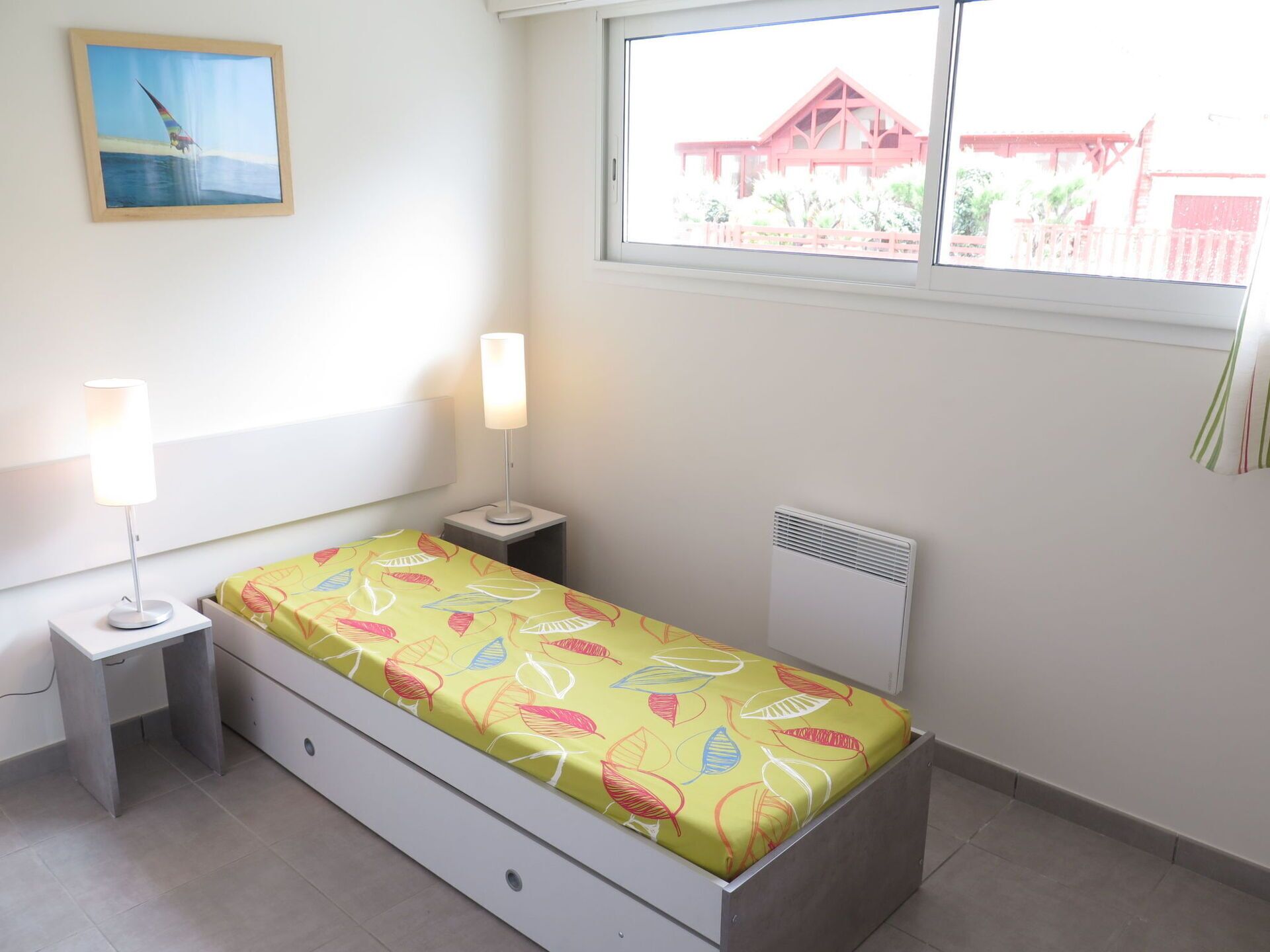 Une petite pièce lumineuse, avec un lit, deux tables de chevet, une fenêtre et un radiateur.
