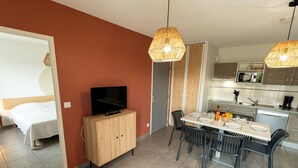 A modern hotel room with a bed, a TV on a wooden cabinet, a dining area with a table and chairs, and a kitchen with a microwave and sink.