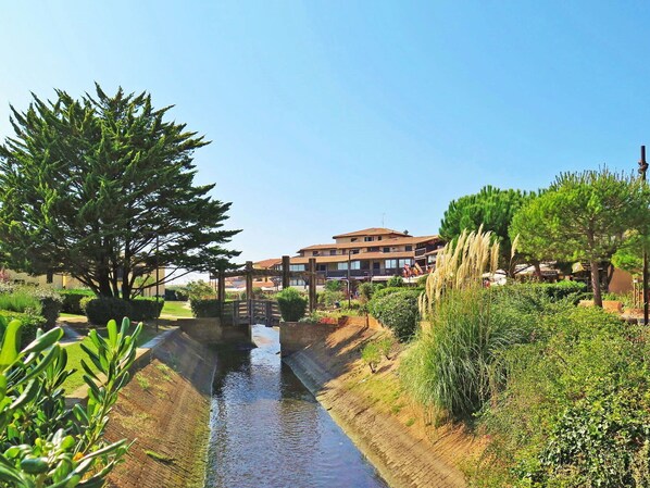 A canal with a wooden bridge, surrounded by greenery and buildings in the background.