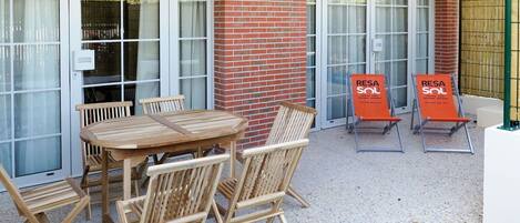 Un espacio al aire libre con mesa y sillas de madera, una pared de ladrillo y un acceso por puerta de cristal.