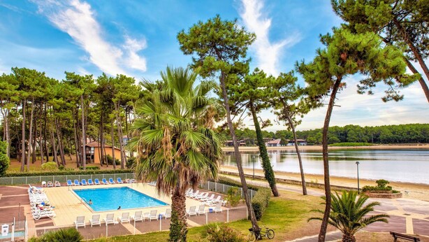 Una piscina con lettini, circondata da palme e pini, con un lago e edifici sullo sfondo.