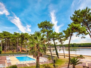 Ein Schwimmbecken mit Liegestühlen, umgeben von Palmen und Kiefern, mit einem See und Gebäuden im Hintergrund.