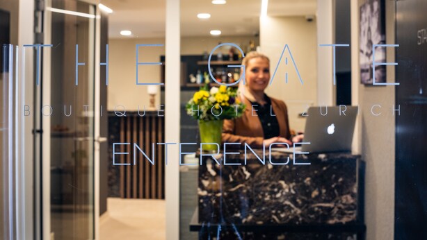 Een hotel receptie met een receptioniste achter de balie, een laptop en een bloevas.