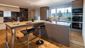 Een moderne keuken met een centraal eiland, barkrukken, roestvrijstalen apparatuur en een groot raam met uitzicht op bomen.