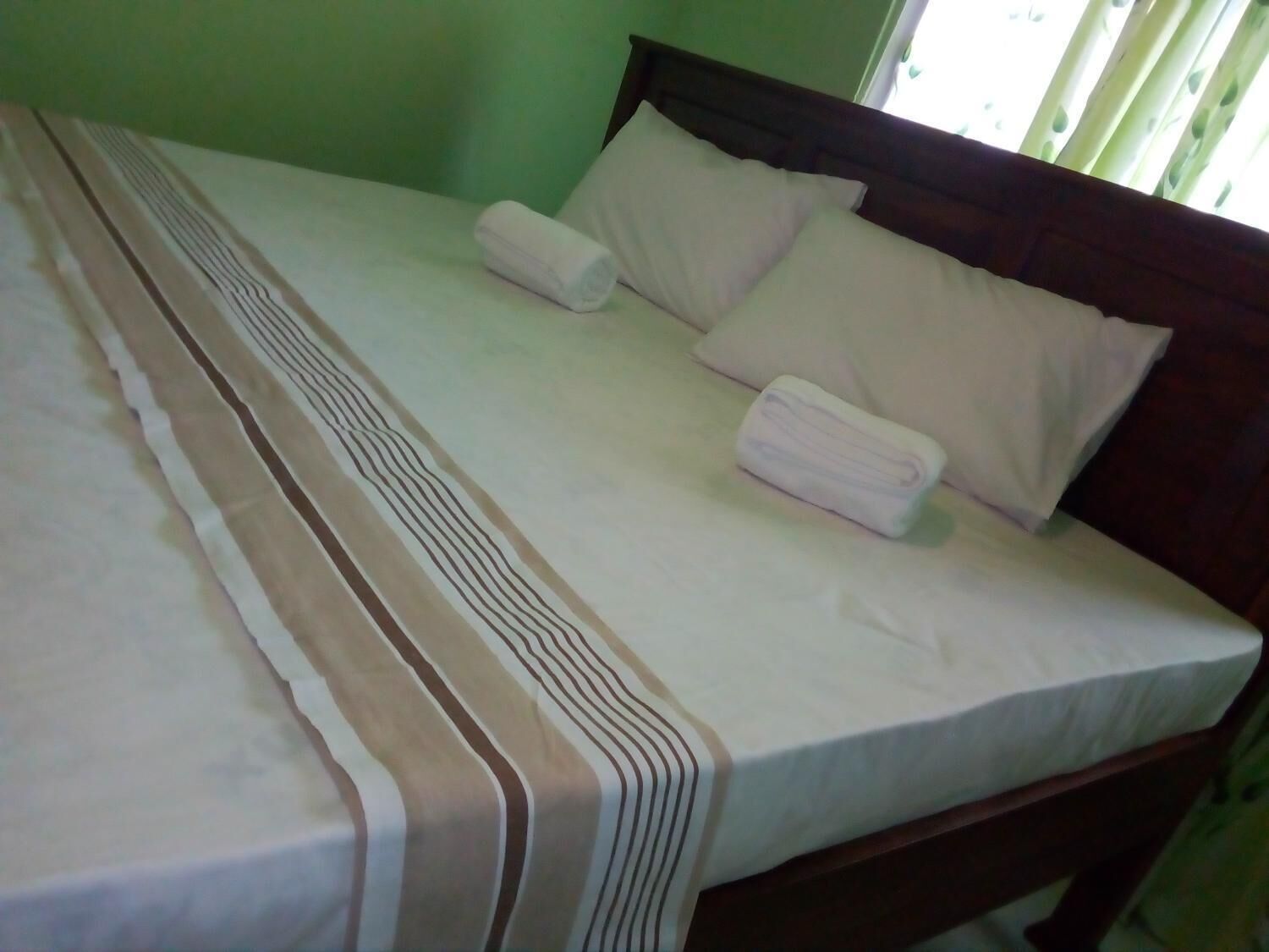 A neatly made bed with a striped bedspread and two pillows, a wooden headboard, and a window with blinds.