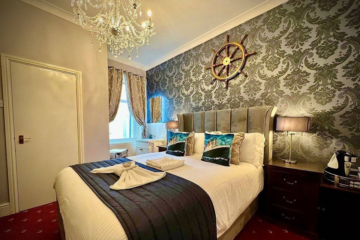 A hotel room with a large bed, a chandelier, a ship's wheel on the wall, and a red carpet.