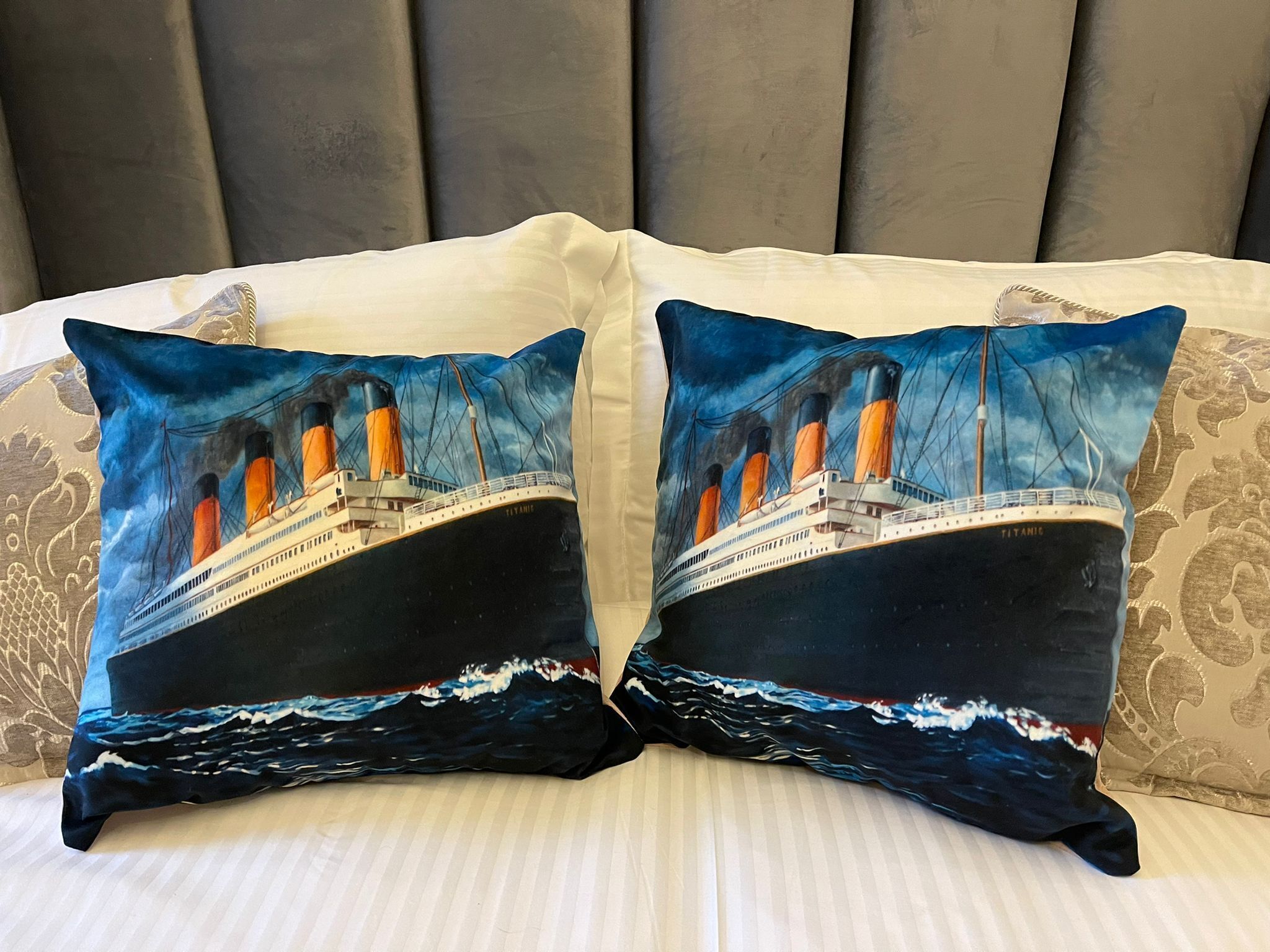 Two pillows with a Titanic ship design on a bed with striped bedding and a grey headboard.