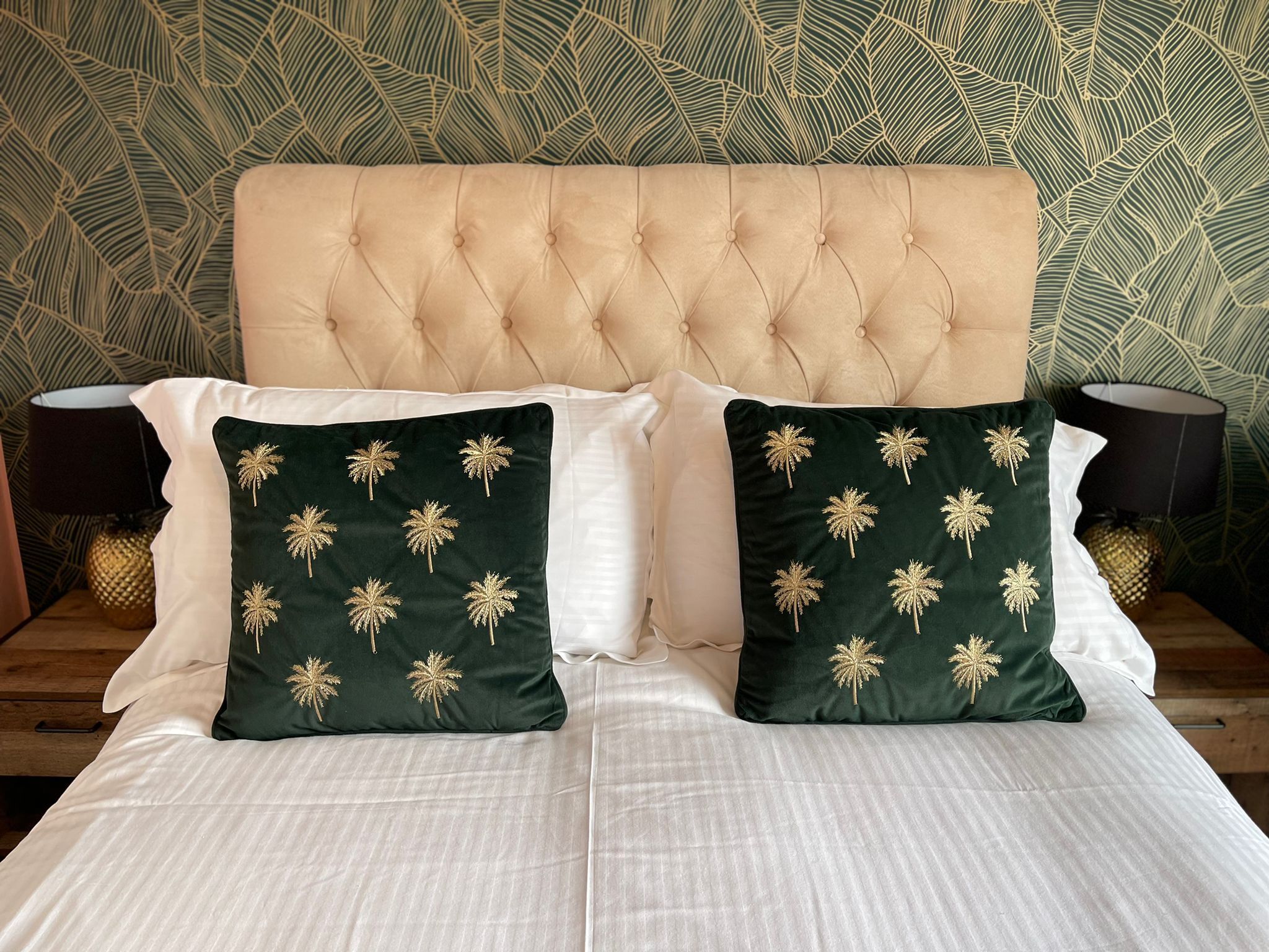 Una cama con cabecera capitonada, dos almohadas verde oscuro con bordado de palmeras doradas y ropa de cama blanca.